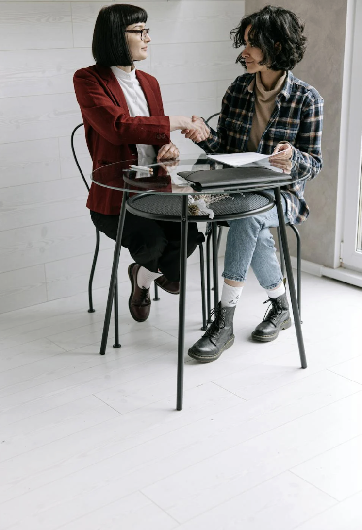 This photo shows two women shaking hands over a desk with documents.